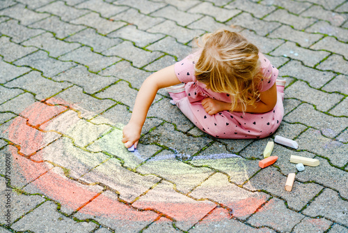 Little preschool girl painting rainbow with colorful chalks on ground on backyard. Positive happy toddler child drawing and creating pictures. Creative outdoors activity in summer. © Irina Schmidt