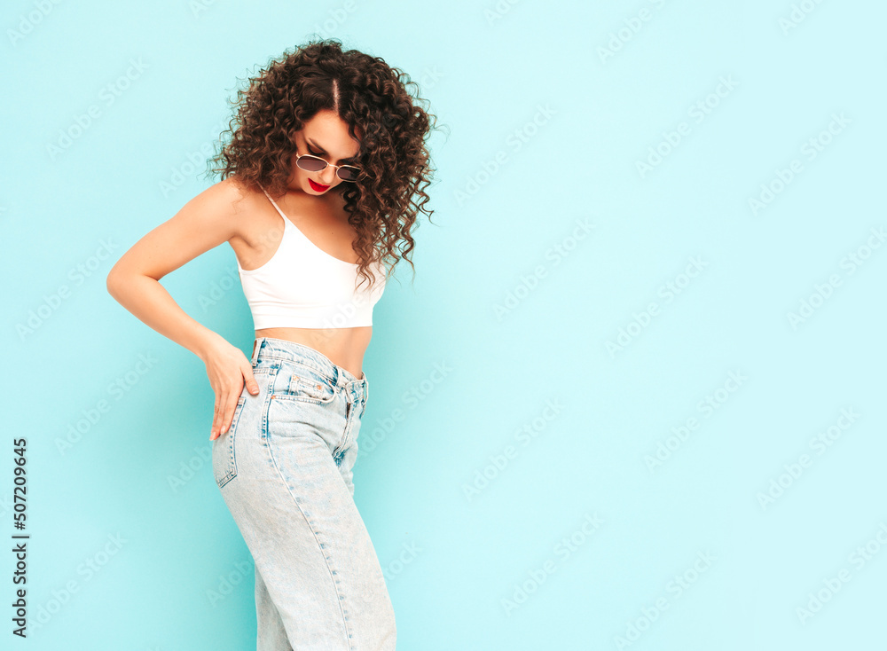 Young beautiful smiling female in trendy summer hipster clothes. carefree woman posing near blue wall in studio. Positive blond model with curls hairstyle. Cheerful and happy. In sunglasses Young beautiful smiling female in trendy summer hipster clothes. carefree woman posing near blue wall in studio. Positive blond model with curls hairstyle. Cheerful and happy. In sunglasses