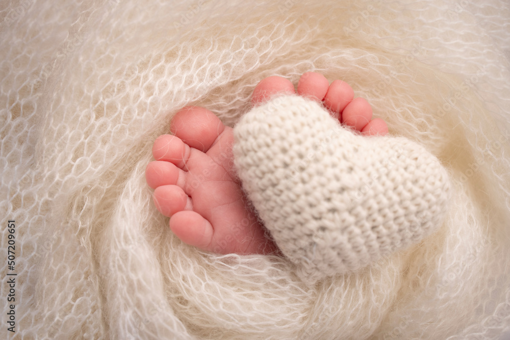 Knitted white heart in the legs of a baby. Soft feet of a new born in a white wool blanket. Close-up of toes, heels and feet of a newborn. Macro photography the tiny foot of a newborn baby. Knitted white heart in the legs of a baby. Soft feet of a new born in a white wool blanket. Close-up of toes, heels and feet of a newborn. Macro photography the tiny foot of a newborn baby.