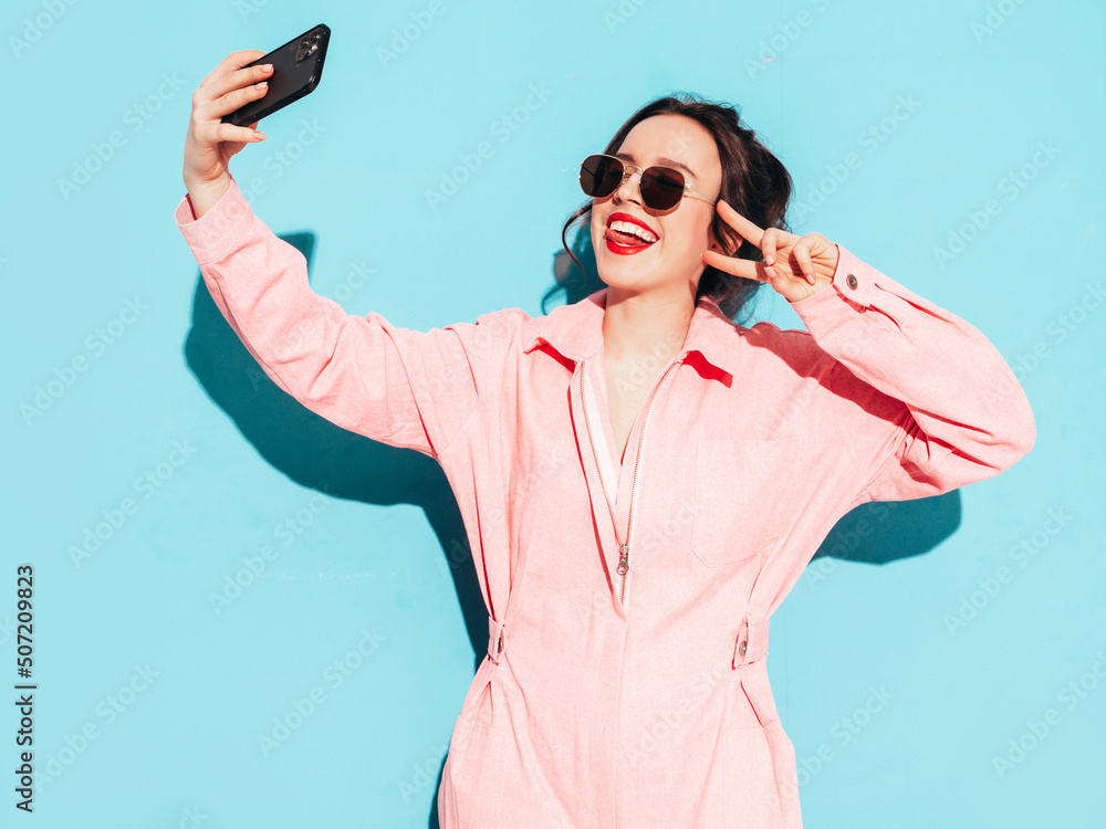 Young beautiful smiling female in trendy summer pink overalls. Sexy carefree woman posing near blue wall in studio with two horns. Positive model having fun and going crazy. Cheerful and happy Young beautiful smiling female in trendy summer pink overalls. Sexy carefree woman posing near blue wall in studio with two horns. Positive model having fun and going crazy. Cheerful and happy