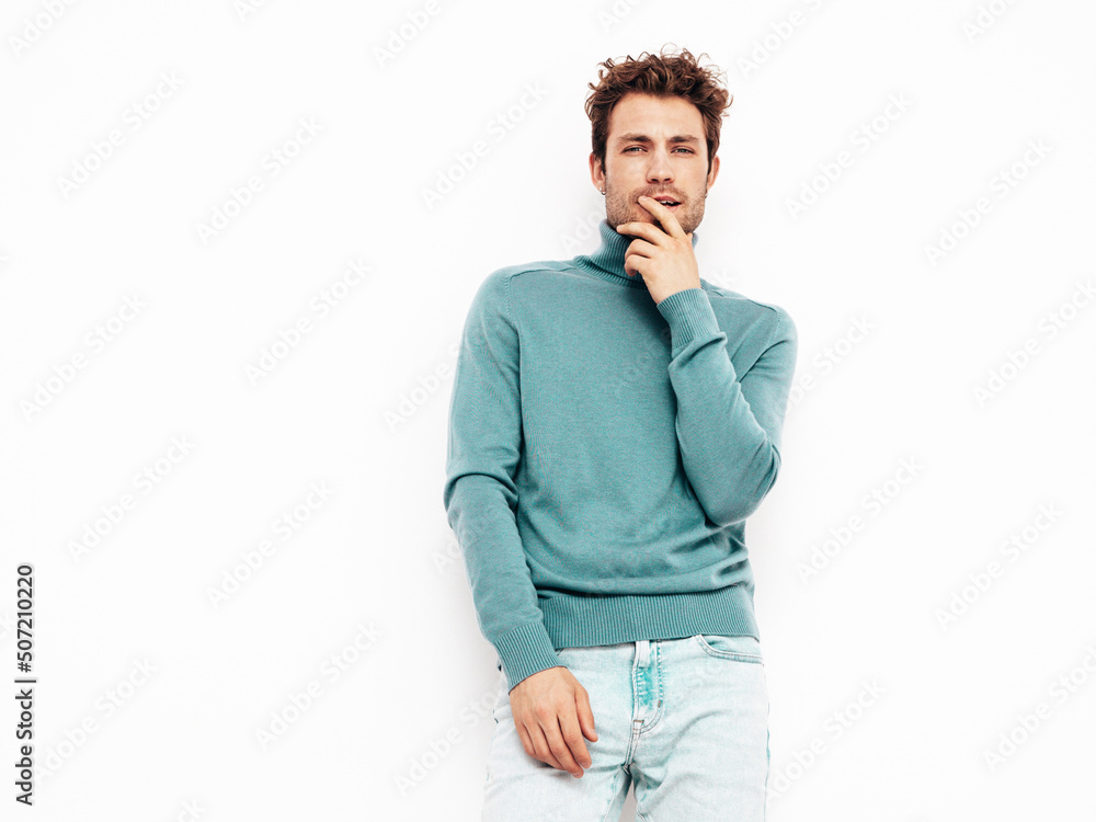 Handsome confident hipster model.Sexy unshaven man dressed in summer stylish blue sweater and jeans clothes. Fashion male with curly hairstyle posing in studio. Isolated on white. Touching his chin Handsome confident hipster model.Sexy unshaven man dressed in summer stylish blue sweater and jeans clothes. Fashion male with curly hairstyle posing in studio. Isolated on white. Touching his chin