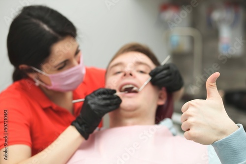 Happy positive satisfied guy show like, thumb up gesture, young man patient is sitting at doctor dentist chair at dental office, woman doctor in medical mask on face. Visiting dentist, check up teeth