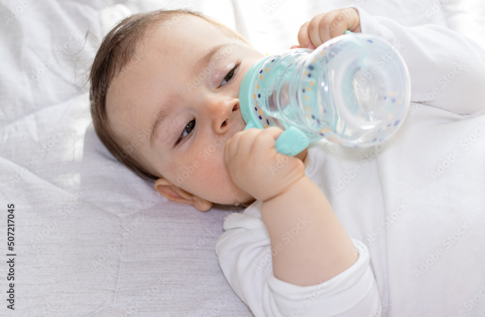 adorable baby boy laying on bed drinking water from plastic bottle with silicone head for mouth. toddler hold legs up. cute baby feet.cup with 2 handles,anti leak, straw wide mouth. adorable baby boy laying on bed drinking water from plastic bottle with silicone head for mouth. toddler hold legs up. cute baby feet.cup with 2 handles,anti leak, straw wide mouth.