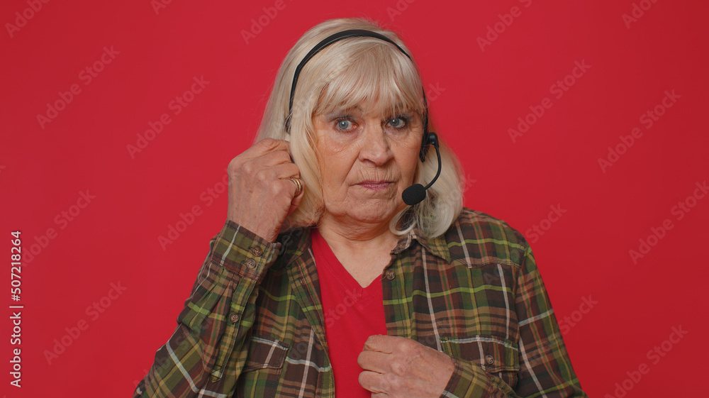 Senior old woman wearing headset, freelance worker, call center or support service operator helpline, having talk with client or colleague, communication support. Elderly grandmother on red background