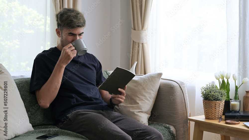 Relaxed millennial man drinking hot coffee and reading book on comfortable sofa