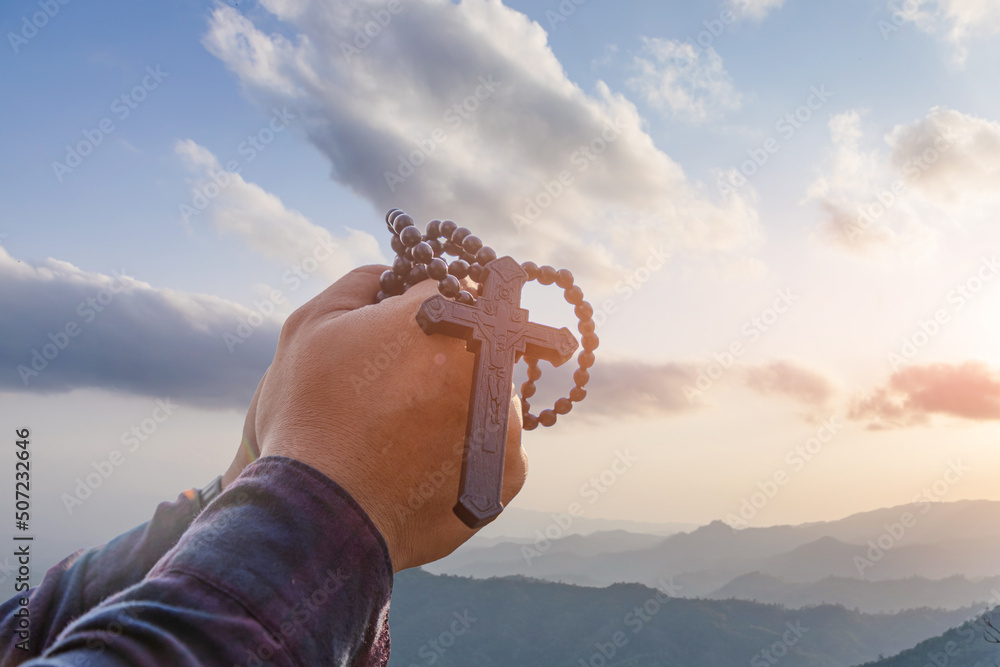 Hands folded holding rosary crucifix symbol with bright sunbeam on the clouds sky background.Religion and Hope Concept. - obrazy, fototapety, plakaty 