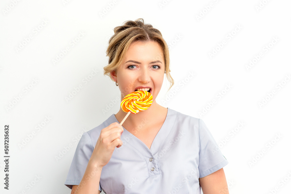 Portrait of a beautiful young caucasian beautician wearing a medical shirt bites a lollipop on a white background Portrait of a beautiful young caucasian beautician wearing a medical shirt bites a lollipop on a white background