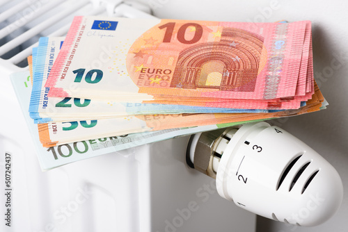 Stack of euro money banknotes on heating radiator battery with thermostat temperature regulator. Concept of expensive heating costs and rising energy bill prices for winter cold season.