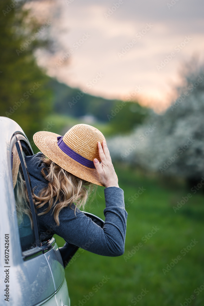 Woman with straw hat looking from car window at sunset. Enjoyment of road trip. Travel on vacation by car