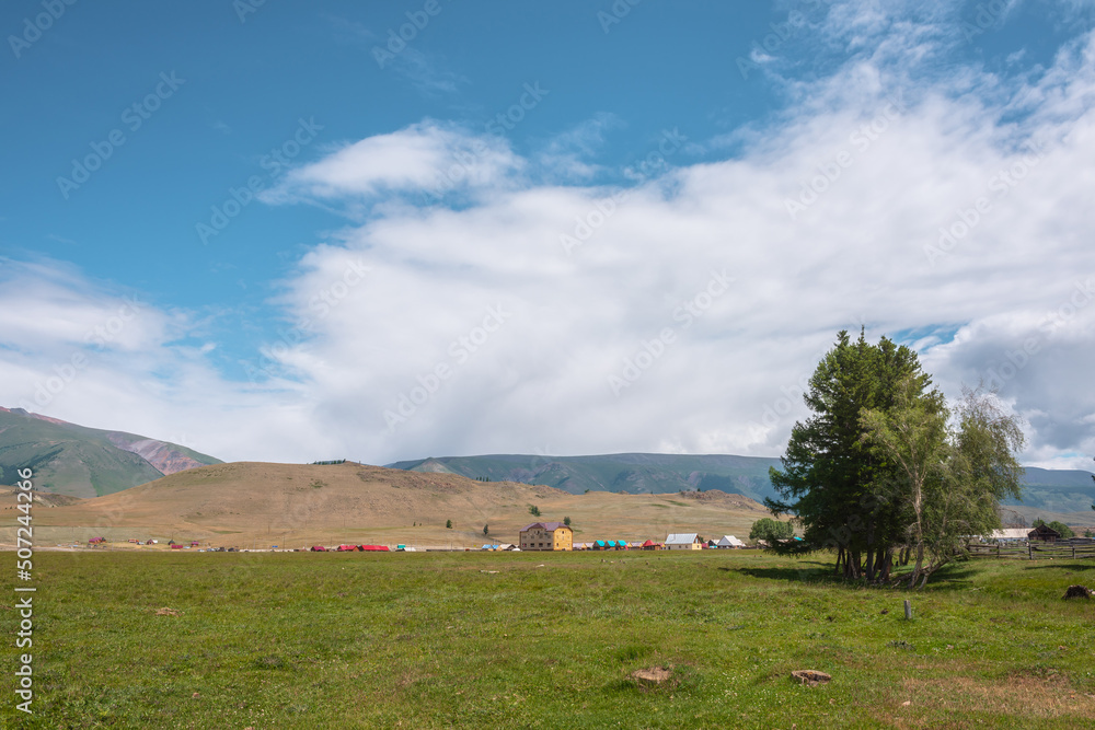 Obraz premium Dramatic alpine view from trees to small houses in sunlit green meadow against high mountain range under clouds in blue sky. Scenic mountain landscape at changeable weather. Village in high mountains.