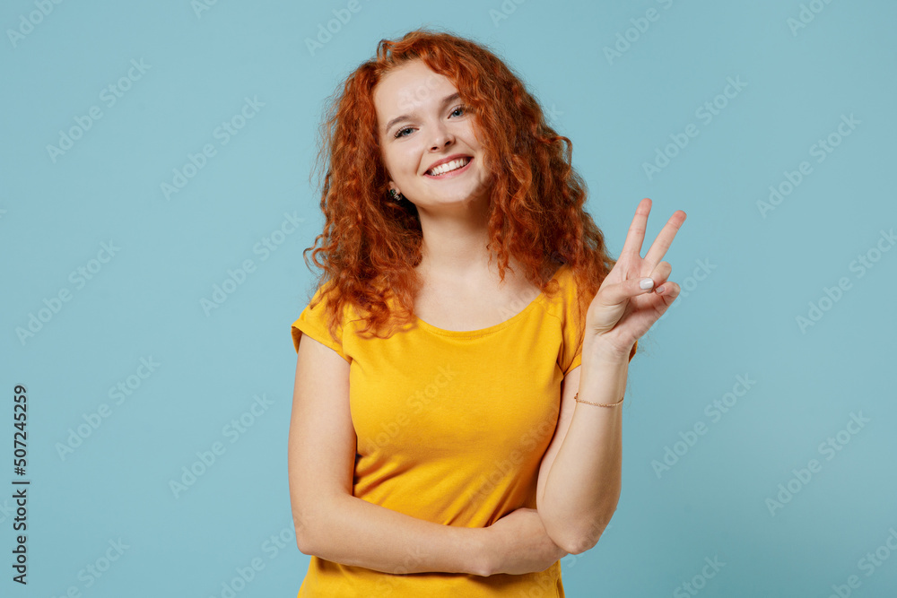 Young smiling happy redhead cheerful friendly woman 20s wear yellow t-shirt look camera showing victory sign isolated on plain light pastel blue background studio portrait. People lifestyle concept Young smiling happy redhead cheerful friendly woman 20s wear yellow t-shirt look camera showing victory sign isolated on plain light pastel blue background studio portrait. People lifestyle concept