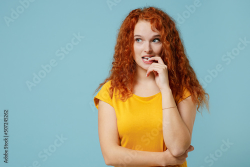 Young thoughtful pensive sad redhead woman 20s wearing yellow t-shirt look aside on workspace area biting nails fingers isolated on plain light pastel blue background studio. People lifestyle concept.