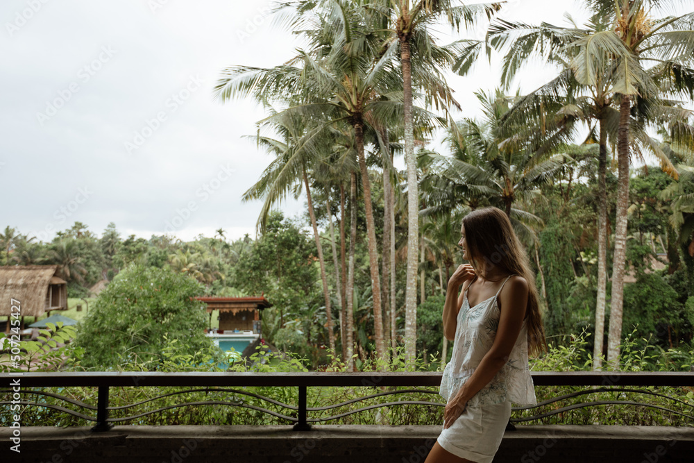 Naklejka premium Young beautiful slender girl stands on the balcony against the backdrop of palm trees