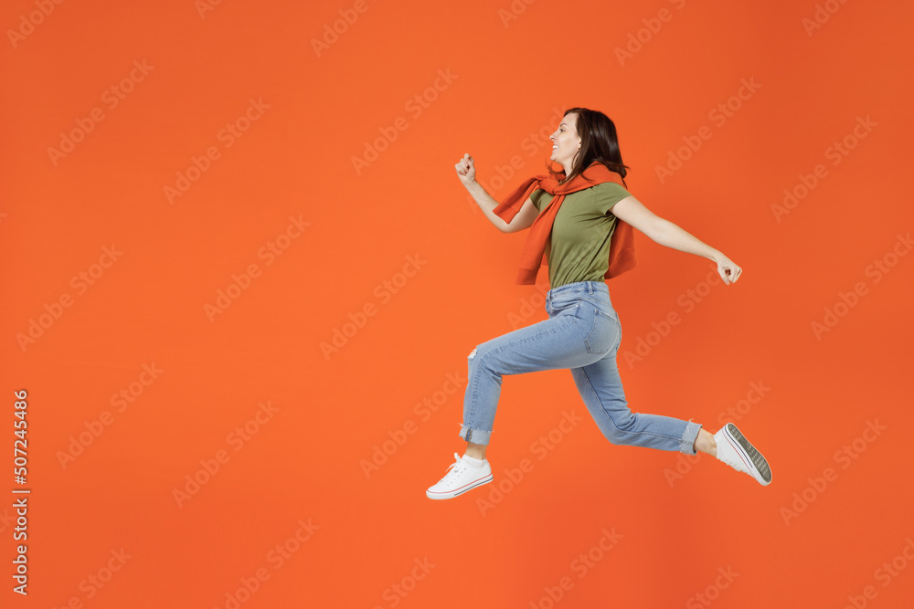 Full body side view young hurrying cool fun happy woman 20s wearing khaki t-shirt tied sweater on shoulders jump high run isolated on plain orange background studio portrait. People lifestyle concept.
