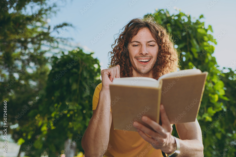 Young surprised satisfied shocked smiling fun cool happy man 20s in yellow t-shirt sitting on bench read book rest relax in spring green city park outdoors on nature. Urban lifestyle leisure concept.