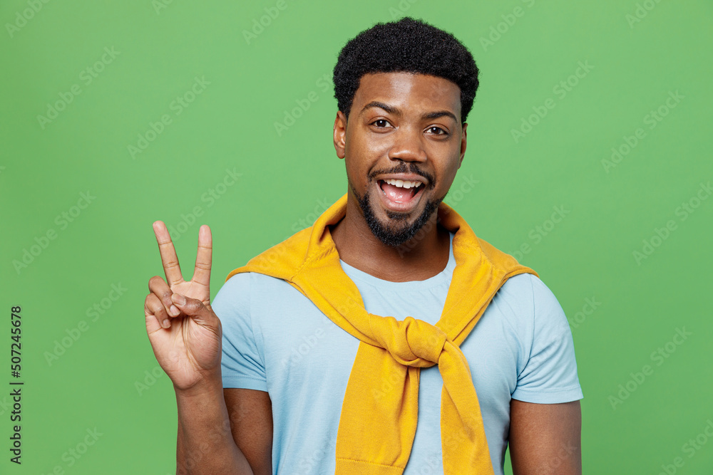 Young smiling happy joyful fun cool satisfied man of African American ethnicity 20s wear blue t-shirt showing victory sign isolated on plain green background studio portrait. People lifestyle concept. Young smiling happy joyful fun cool satisfied man of African American ethnicity 20s wear blue t-shirt showing victory sign isolated on plain green background studio portrait. People lifestyle concept.