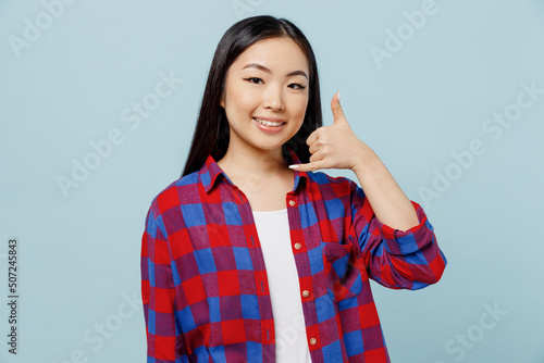 Young smiling cheerful fun woman of Asian ethnicity 20s wear checkered shirt doing phone gesture like says call me back isolated on plain pastel light blue color background. People lifestyle concept.