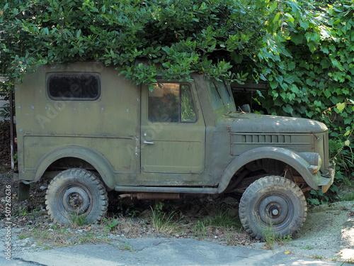 An old khaki-colored truck is parked on the side of a broken rural road in a thicket of green bushes
