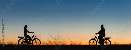 An elderly man, a pensioner and a girl on electric bikes ride towards each other against the backdrop of the sunset sky. Silhouettes of people in profile. Active retirement, travel and sports.