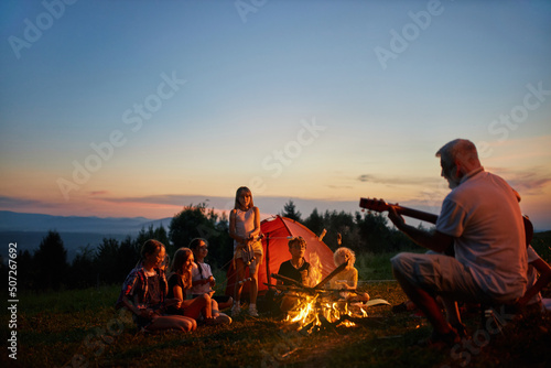 Happy children sitting around campfire, singing songs with guitar at dusk. Side view adult guide playing guitar for kids near fire in mountains, with colorful sky on background. Concept of camping.
