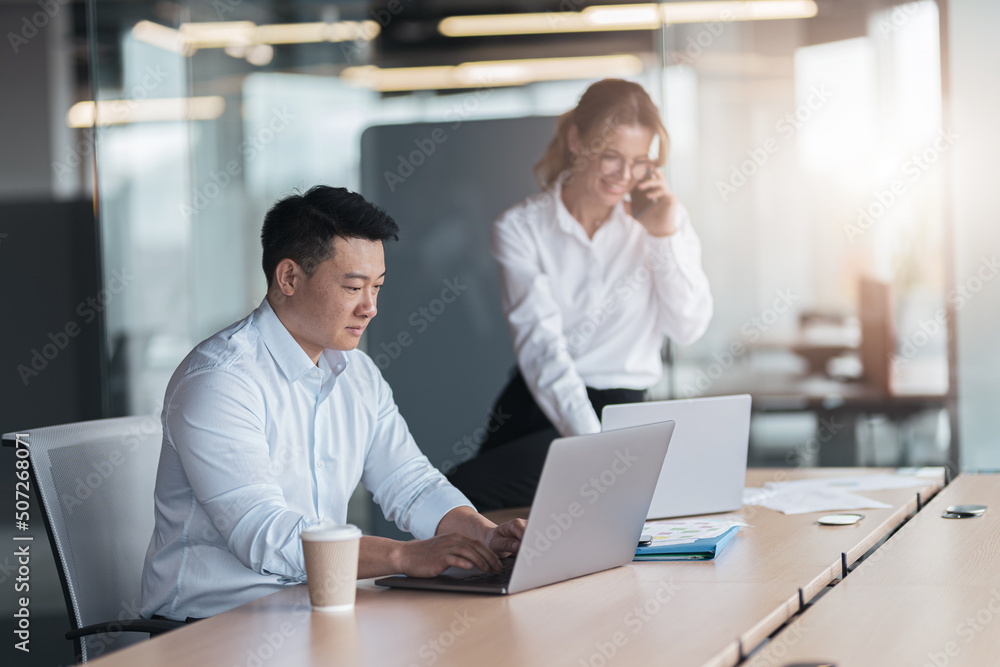 Asian businessman working on laptop with colleague talking on phone in background Asian businessman working on laptop with colleague talking on phone in background