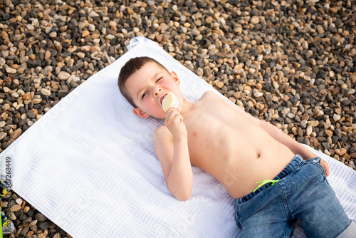 Cute six year old boy eating white chocolate ice cream on a stick while lying on a white towel on the rocks photo