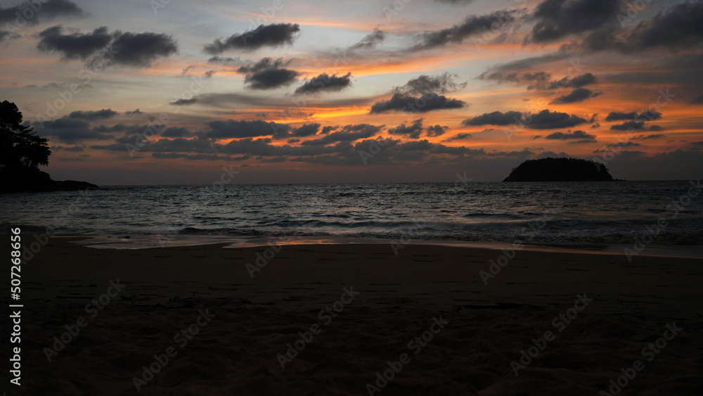 An epic sunset with a view of the sea and the island. The orange sky is covered with dark clouds. There is a lonely island in sea. Silhouettes of trees are visible. The sky is reflected on wet sand