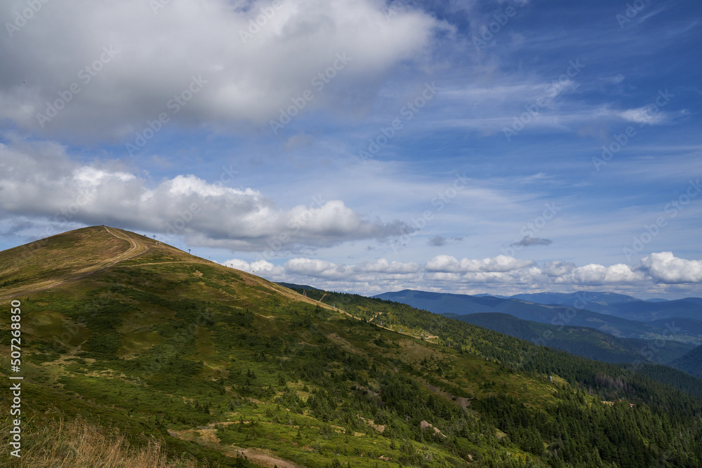 Fototapeta premium Scenic view of gentle slope mountain, with path leading to hilltop in summer. Cumulus clouds, crossing blue sky, seemed touching top of mount, with hilly landscape on background. Concept of highland.