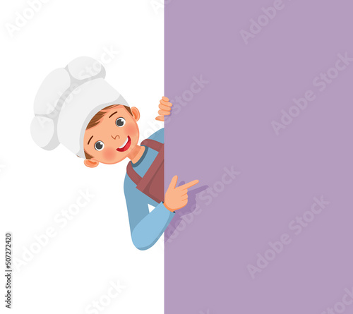 cute little boy in chef hat and apron peeking from board pointing finger to empty banner for copy space