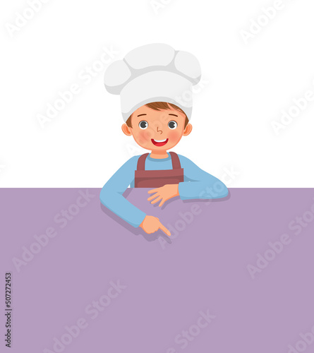 cute little boy in chef hat and apron standing behind empty board pointing finger to blank banner for copy space