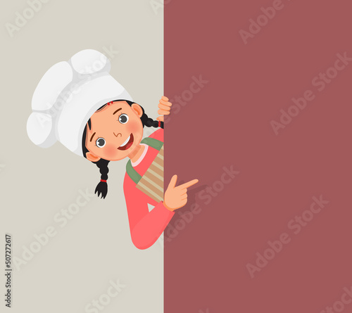 cute little girl in chef hat and apron peeking from board pointing finger to empty banner for copy space