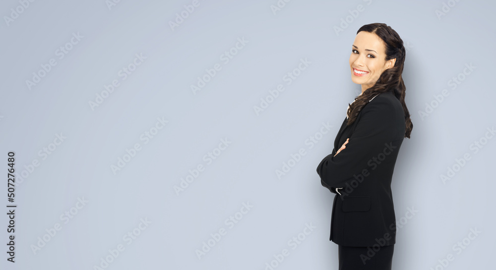 Happy smiling attractive businesswoman, with copy space area for some slogan or advertising text. Confident brunette woman in black suit standing against grey background. Business ad concept. Happy smiling attractive businesswoman, with copy space area for some slogan or advertising text. Confident brunette woman in black suit standing against grey background. Business ad concept.