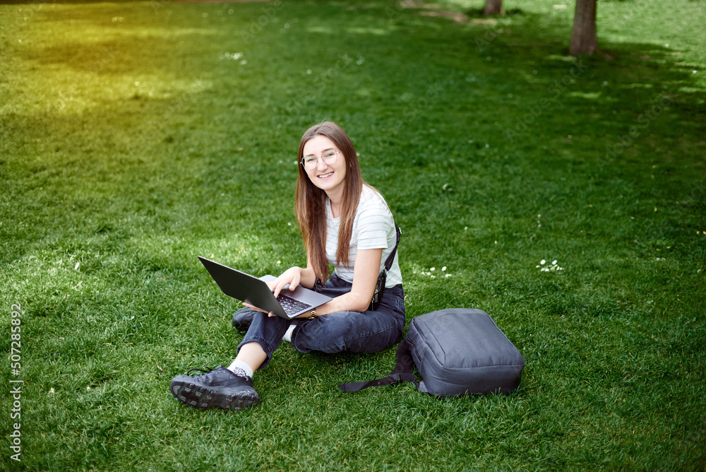 A young girl works with a laptop in the fresh air in the park, sitting on the lawn. The girl takes courses on a laptop and smiles. The woman looks into the camera while sitting on the lawn. A young girl works with a laptop in the fresh air in the park, sitting on the lawn. The girl takes courses on a laptop and smiles. The woman looks into the camera while sitting on the lawn.
