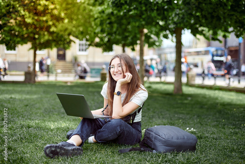 A woman sits on the lawn with a laptop dreaming. A woman works remotely with a laptop outdoors in the park. The concept of remote work. A happy girl takes courses.