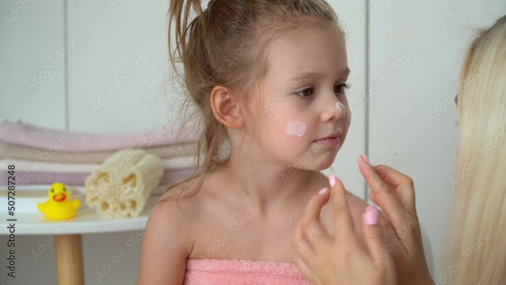 Mother puts cream to little daughters skin face in bathroom. Happy child after bathing. Baby care, moisturizer. Spa, clean, hygiene, health Mother puts cream to little daughters skin face in bathroom. Happy child after bathing. Baby care, moisturizer. Spa, clean, hygiene, health