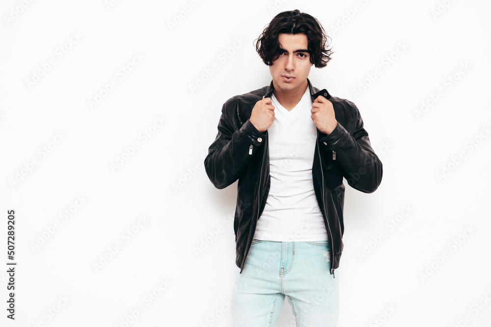 Portrait of handsome confident stylish hipster lambersexual model.Sexy man dressed in black leather biker jacket and jeans. Fashion male isolated in studio. Posing near white wall Portrait of handsome confident stylish hipster lambersexual model.Sexy man dressed in black leather biker jacket and jeans. Fashion male isolated in studio. Posing near white wall