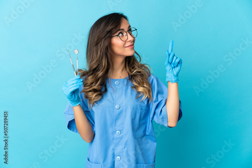 Woman dentist holding tools over isolated on blue background pointing up a great idea