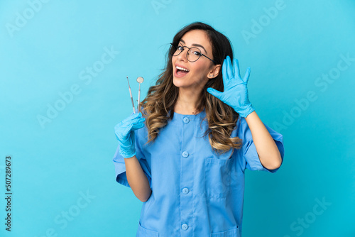 Woman dentist holding tools over isolated on blue background listening to something by putting hand on the ear