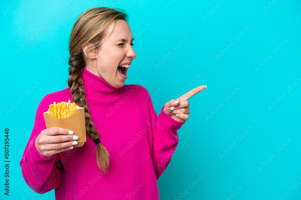 Young caucasian woman holding fried chips isolated on blue background pointing finger to the side and presenting a product Young caucasian woman holding fried chips isolated on blue background pointing finger to the side and presenting a product