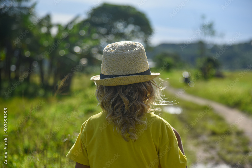 A little curly-haired boy walks in nature in a hat and a yellow T-shirt. Family vacation, traveler, trip, summer time. Green grass in the background, back view