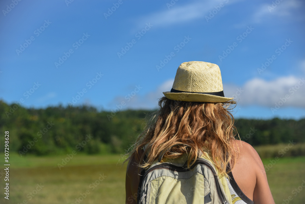 A curly-haired man walks in nature in a hat and with backpack. Vacation, traveler, trip, summer time. Green grass and blue sky in the background, lifestyle, back view