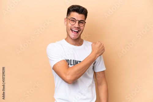 Young volunteer caucasian man isolated on beige background celebrating a victory