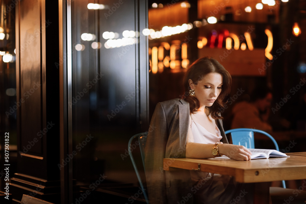 portrait of young beautiful woman is reading paper book in cafe in dusk. girl is online study, remote work. portrait of young beautiful woman is reading paper book in cafe in dusk. girl is online study, remote work.