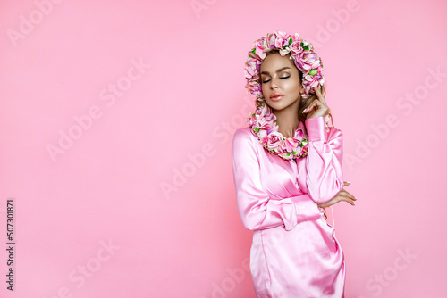 Spring fashion model. Beautiful natural woman is wearing pink dress and wreath with flowers. Elegant woman is holding bouquet on pink background. Studio shoot. © marcink3333