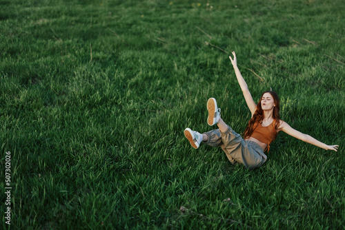A young woman playing games in the park on the green grass spreading her arms and legs in different directions falling and smiling in the summer sunlight