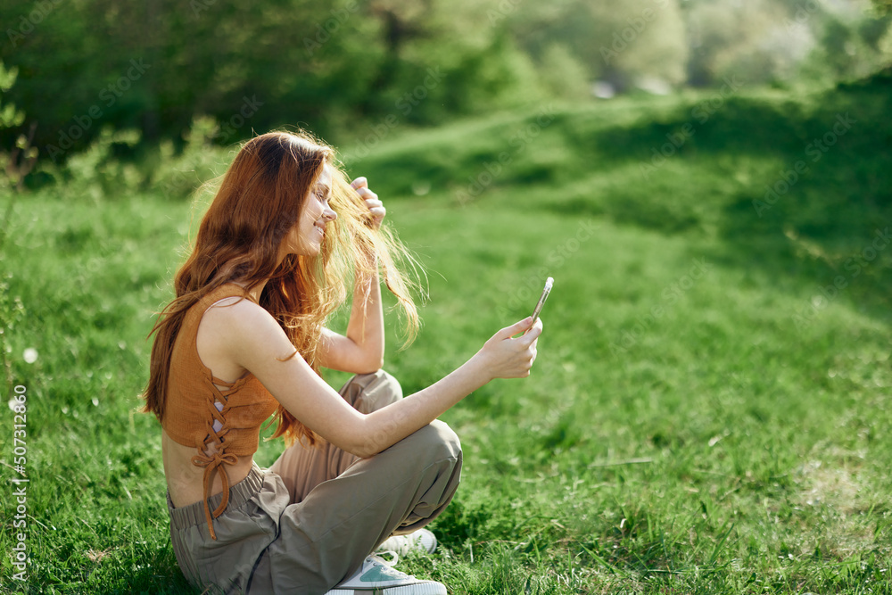 Fototapeta premium A young happy woman sitting in the park on the green grass in nature with her phone in her hands in her casual clothes and chatting via video link, the concept of healthy recreation meditation 