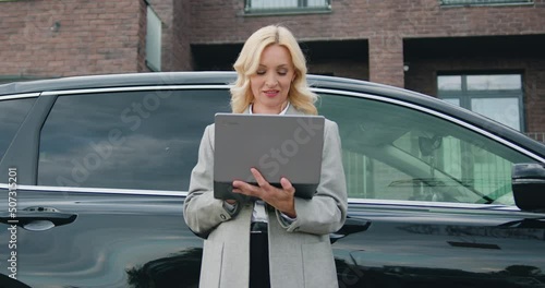 Attractive Blonde female driver standing near her car using laptop on a city street. Smiling while reading new messages or browse videos or photos or social networks