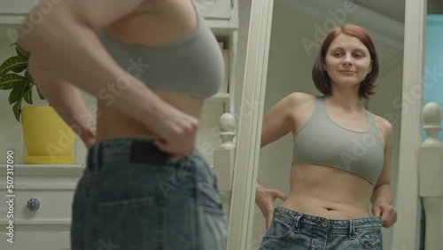 30s woman looking at mirror for result of weight loss. Female wearing oversized jeans because of weight loss.