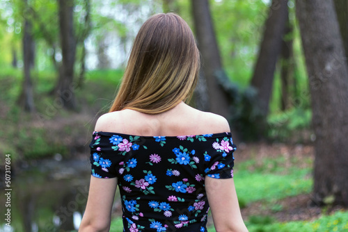 young European girl with long hair, dressed in a bright off-the-shoulder dress, walks through the spring park, back view from behind