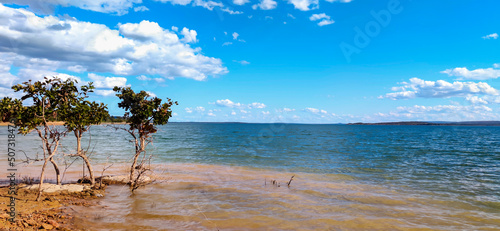Linda vista de parte da represa de três marias, com água límpida, céu azul com nuvens e pequenas árvores na beirada, na região de Três Marias, Minas Gerais, Brasil.